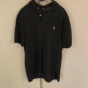 Svart pikétröja från Ralph Lauren - Snygg svart pikétröja från Ralph Lauren med klassisk passform. Tröjan har korta ärmar och en liten broderad logotyp i rött på bröstet. Perfekt för en stilren look.