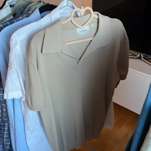 Pikétröja från Sandö - Säljer en stilren beige/kaki pikétröja från Sandö. Tröjan har korta ärmar och en klassisk krage. 