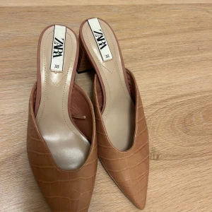 Bruna klackskor från Zara - Snygga bruna klackskor från Zara med krokodilpräglat mönster och spetsig tå. Skorna har en bred klack som ger stabilitet och stil. Perfekta för att lyfta din outfit med en touch av elegans.