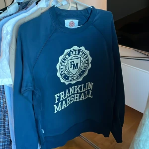 Vintage sweatshirt från Franklin & Marshall - Säljer en blå sweatshirt från Franklin & Marshall med tryck på framsidan. Tröjan har långa ärmar och en rund halsringning. Perfekt för en avslappnad stil.