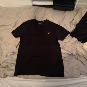 Svart t-shirt från Lyle & Scott - Snygg svart t-shirt från Lyle & Scott med deras ikoniska gula logga på bröstet. Perfekt för en stilren och avslappnad look. Passar till både jeans och shorts.