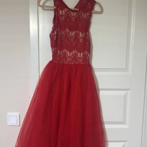 Röd spetsklänning med öppen rygg - Elegant röd klänning med spetsdetaljer och öppen rygg. Den har en vacker, lång kjol och är perfekt för speciella tillfällen. Klänningen har en ärmlös design och en fin passform.