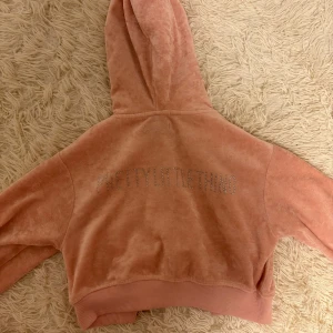 Rosa hoodie från PrettyLittleThing - Säljer en rosa hoodie från PrettyLittleThing med glittrande text på ryggen. Hoodien är croppad och har en dragkedja framtill. Perfekt för en avslappnad stil.