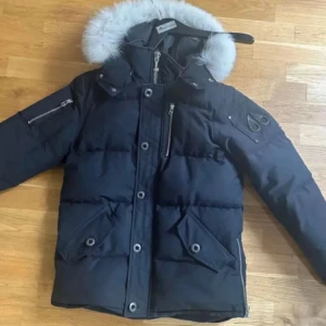 Svart dunjacka med pälsdetalj - Snygg svart dunjacka från Woolrich med avtagbar huva och pälsdetalj. Jackan har flera praktiska fickor med knappar och dragkedja. Perfekt för kalla dagar.