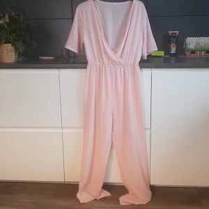 Ljusrosa omlott jumpsuit - Säljer en ljusrosa jumpsuit med omlott-design och korta ärmar. Den har en v-ringad hals och elastisk midja för en bekväm passform. Perfekt för en avslappnad och stilren look.