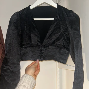 Svart topp - Snygg svart silkes topp med knappar framtill. Den har en croppad passform och långa ärmar, perfekt för en festlig look.