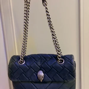 Svart quiltad axelväska från The Kensington - Snygg svart quiltad axelväska med silverkedja och dekorativt spänne framtill. Säljer billigt pågrund av att den är sliten.