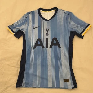 Tottenham fotbollströja från Nike 24/25 - Snygg blå randig fotbollströja från Nike med Tottenham Hotspur-logga och AIA sponsortryck. Tröjan har korta ärmar med gula detaljer och Premier League-märke på ärmen. Perfekt för fans av Spurs!   Mer bilder kan fås och pris kan diskuteras!!