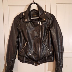 Svart "skinnjacka" från Giorgia Fashion - Snygg svart skinnjacka från Giorgia Fashion i storlek M. Jackan har en klassisk bikerstil med dragkedja och knappar, samt ett bälte i midjan. Tillverkad i polyuretan med polyester- och viskosfoder.                                      Jackan är sparsamt använd och i mycket bra skick.