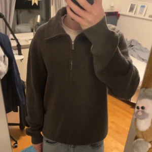 Grå stickad tröja från Bläck - Säljer en stilren grå stickad tröja från Bläck med dragkedja och hög krage. Perfekt för kyliga dagar med sin ribbade design och långa ärmar. Passar både till jeans och chinos.