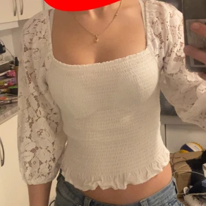 Vit blus med spetsdetaljer - Säljer en söt vit blus med spetsdetaljer och trekvartsärmar. Blusen har en smockad kropp och volangkant nedtill, vilket ger en romantisk touch. Perfekt för en somrig look!