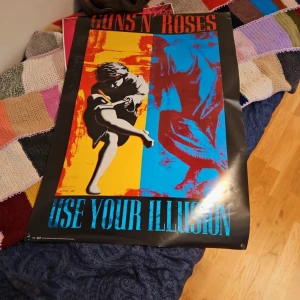 Guns N' Roses Use Your Illusion affisch - Färgglad affisch med motiv från Guns N' Roses album 'Use Your Illusion'. Designen har starka kontraster i gult, rött och blått med en ikonisk bild av en ängel. Perfekt för musikälskare och samlare. 