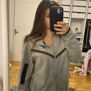 Grå jacka från Nike - Säljer en grå jacka från Nike med dragkedja och svarta detaljer. Jackan har långa ärmar och en normal passform. Perfekt för en sportig look. Inga fläckar. Äkta 