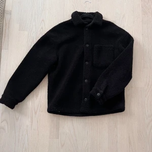 Svart teddyjacka från Zara - Säljer en svart teddyjacka från Zara i storlek M. Jackan har en avslappnad passform med knappar framtill och en bröstficka. Perfekt för kyligare dagar.