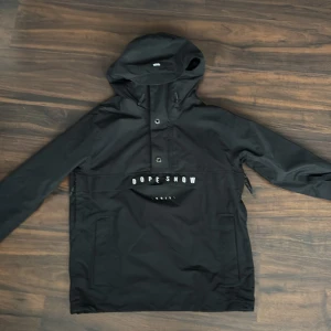 Svart jacka från Dope Snow - Snygg svart jacka från Dope Snow med huva och dragkedja. Finns 2 fickor på magen och en ficka på armen för liftkort. Finns även dragkedja på sidorna för att öppna upp jackan om det blir för varmt! Nypris: 1899kr