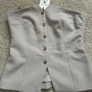 Beige korsettopp med knappar - Elegant beige korsettopp med knappar framtill. Toppen har en stilren design med en liten slits nedtill och är perfekt för en chic look.