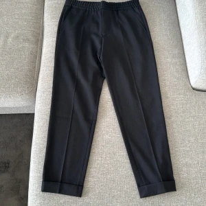 Svarta byxor från Filippa K - Snygga svarta byxor från Filippa K med elastisk midja och raka ben. Byxorna har diskreta bakfickor och är perfekta för en stilren look. Passar både till vardags och mer uppklädda tillfällen.