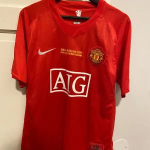 Röd Manchester United fotbollströja - Säljer en unik röd Manchester United fotbollströja med design från 2008 med AIG-logga på framsidan. Tröjan har korta ärmar och ett broderat klubbmärke. Texten 'Final Moscow 2008' är tryckt ovanför loggan. Perfekt för fans av laget! Varan är ej äkta! Hör av er om ni har fler frågor.