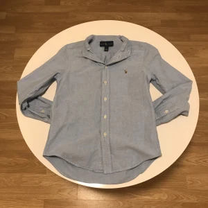 Blå skjorta från Ralph Lauren - Snygg ljusblå skjorta från Ralph Lauren med klassisk krage och broderad logotyp på bröstet. Skjortan har långa ärmar och knappar framtill. Perfekt för en stilren look.