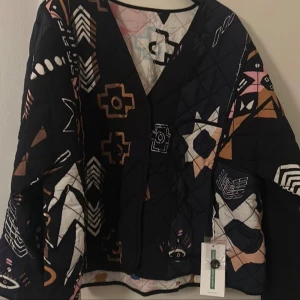 Munthe jacka - En snygg och unik quiltad jacka från Munthe. Jackan har ett vackert mönster med geometriska former och stjärnor i olika färger. Den har en v-ringning och en knappstängning framtill. Jackan är perfekt för att addera en touch av stil till din outfit.vändbar