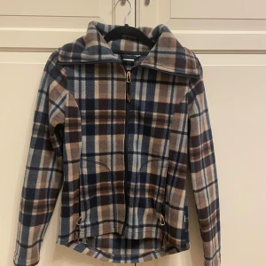 Rutig fleecejacka från CRW Hööks  - Säljer en mysig rutig fleecejacka från CRW i blå, brun och beige. Jackan har en dragkedja framtill och långa ärmar. Perfekt för kyliga dagar!