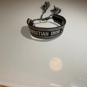 Svart och vit armband från Christian Dior - Snyggt armband från Christian Dior i svart och vitt med texten 'J'ADIOR'. Armbandet har justerbara tofsar för perfekt passform. Perfekt accessoar för att ge din stil en lyxig touch.