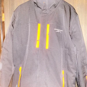 Svart jacka från Swedemount - Säljer en svart jacka från Swedemount med orange detaljer och dragkedjor. Jackan har en hög krage och flera praktiska fickor. Perfekt för utomhusaktiviteter. Aldrig använd 