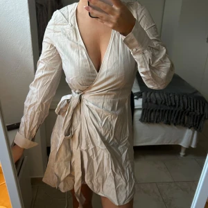 Beige omlottklänning - Snygg beige omlottklänning med knytband i midjan och v-ringad hals. Klänningen har långa ärmar och en kort längd, perfekt för en stilren look.