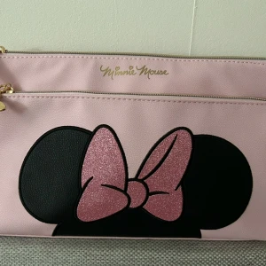 Äkta Rosa Minnie Mouse kuvertväska från Disney - Söt rosa kuvertväska med ett glittrigt motiv av Minnie Mouse. Väskan har en dragkedja upptill och är perfekt för att hålla dina småsaker organiserade. Den är tillverkad i syntetmaterial och har en elegant design med guldfärgade detaljer.