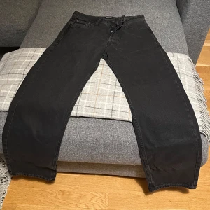 Svarta jeans från Jack & Jones - Säljer ett par helt nya svarta jeans från Jack & Jones i modellen Chris. De har en avslappnad passform och är perfekta för en casual look. Jeansen har en klassisk design med knappar och dragkedja. Obs - de är inte stulna, de kom i fel storlek och vi hann inte returnera dem innan tiden var ute. Nypris 499:-  