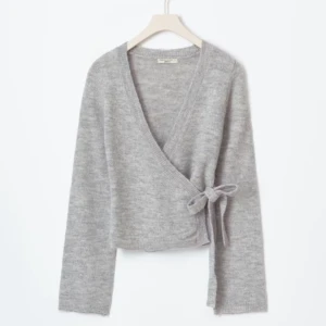 Grå omlottkofta - Säljer en mysig grå omlottkofta med knytband i sidan. Koftan har långa ärmar och är perfekt för kyligare dagar. Den är tillverkad i ett mjukt material som ger en skön känsla. Passar både till jeans och kjol. Storlek M men passar S också!