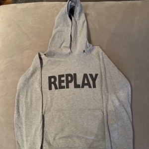 Grå hoodie från Replay - Säljer en stilren grå hoodie från Replay med svart text på framsidan. Tröjan har en klassisk design med huva och långa ärmar, perfekt för en avslappnad look.