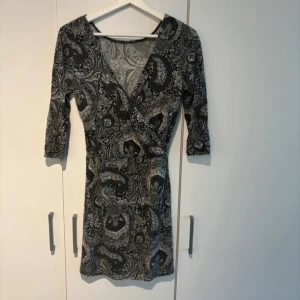 Mönstrad klänning med trekvartsärm - Säljer en snygg klänning med ett unikt mönster i svart och grått. Den har en omlott-design framtill och trekvartsärmar. Perfekt för både vardag och fest!