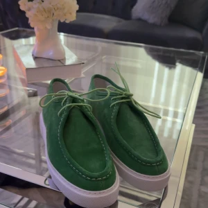 Gröna mockaskor med vit sula - Säljer ett par snygga gröna mockaskor med vit sula. De har en stilren design med snöre och är perfekta för både vardag och fest. Skorna är i mycket bra skick och passar perfekt till vår och sommar. Storleken är normal och de är superbekväma att gå i. Perfekt för dig som vill sticka ut lite! 🌿