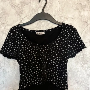 Svart blommig topp från new Yorker  - Säljer en söt svart topp från new Yorker med vita blommor. Den har korta ärmar och en knäppning framtill. Perfekt för sommardagar eller en avslappnad vårlook. Toppen är i bra skick och passar till både jeans och kjol.
