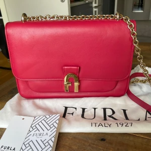 Furla axelremsväska  - Jättefin Furla väska som endast används vid ett tillfälle,så toppen skick. Längd 26 cm,höjd 18 cm. Läder.Färg hallonröd.