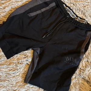 Svarta och grå shorts från Boss - Säljer ett par snygga shorts från Boss i svart och grått. De har en elastisk midja med dragsko och en diskret logga på benet. Perfekta för en avslappnad stil eller träning. Materialet känns mjukt och bekvämt, och de har praktiska fickor på sidorna.