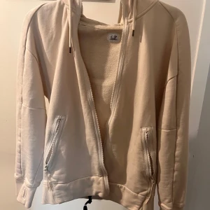 Beige hoodie från C.P. - Säljer en snygg beige hoodie från C.P. med dragkedja och huva. Perfekt för både vår och höst. Den har långa ärmar och praktiska fickor med dragkedjor. Superbekväm och stilren för en casual look.