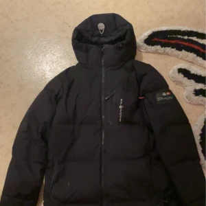 Svart dunjacka från Sail racinga  - Säljer en svart dunjacka från   Sail racinga i mycket bra skick. Den är perfekt för kalla vinterdagar med sin varma vaddering och stilrena design. Jackan har en dragkedja framtill och en hög krage för extra skydd mot kyla. Passar både till vardags och utomhusaktiviteter. Kan pruta vid snabb affär 