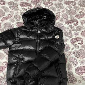 Svart dunjacka från Moncler - Säljer en snygg och varm svart dunjacka från Moncler. Jackan har en glansig finish och är perfekt för kalla dagar. Den har en dragkedja framtill och praktiska fickor med dragkedjor. Moncler-loggan syns tydligt på ärmen. Perfekt för vintern! ❄️ jackan är inte använd den är helt ny. Och den är i storlek 1