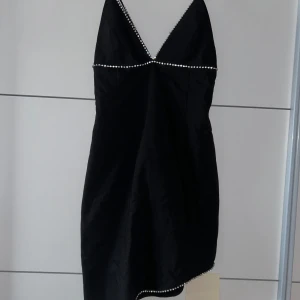 Svart asymmetrisk klänning från Adoore - Snygg svart asymmetrisk klänning från Adoore med glittriga detaljer längs halsringningen. Perfekt för festliga tillfällen! Klänningen har en elegant passform och smala axelband som ger en stilren look. Den är tillverkad i ett mjukt material som känns skönt mot huden. Perfekt till nyår. Kan mötas upp nära Malmö, liten i storleken så passar 40-42 