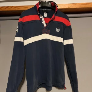 Mörkblå pikétröja från Sail Racing - Säljer en snygg mörkblå pikétröja från Sail Racing med röda och vita detaljer. Tröjan har långa ärmar och knappar vid kragen. Perfekt för en sportig look eller en avslappnad dag ute. Stor logga och nummer 77 på ärmen ger en cool touch. Passar bra till både jeans och shorts!