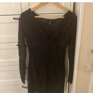 Svart spetsklänning - Snygg svart spetsklänning i mycket bra skick. Den har en v-ringad design och långa ärmar, perfekt för festliga tillfällen. Klänningen är i en kort modell och har en snygg spetsdetalj.