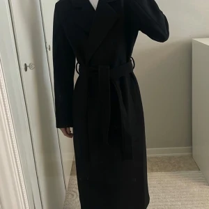 Svart kappa med bälte - Säljer en elegant svart kappa i mycket bra skick. Den har ett klassiskt snitt med ett bälte i midjan för en snygg siluett. Perfekt för höst och vinter.