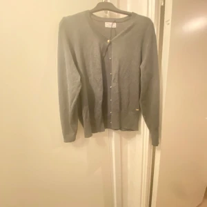 Grå kofta från Holly & Whyte - Säljer en stilren grå kofta från Holly & Whyte. Den har långa ärmar och knappar framtill, perfekt för både vardag och lite finare tillfällen. Materialet känns mjukt och bekvämt, vilket gör den idealisk för kyligare dagar. Passar bra till både jeans och kjol!  Priset kan diskuteras!