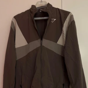 Brun vindjacka från Gymshark - Säljer en brun vindjacka från Gymshark med grå och vita detaljer. Jackan har dragkedja framtill och långa ärmar. Perfekt för träning eller som en lätt jacka till vardags. Gymshark-loggan syns tydligt på ryggen. Nyskick.