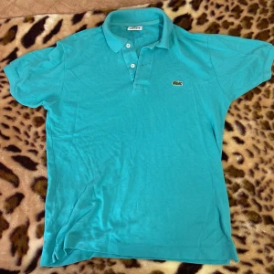Turkos pikétröja från Lacoste - Säljer en snygg turkos pikétröja från Lacoste. Den har den klassiska krokodilloggan på bröstet och tre knappar vid halsen. Perfekt för en avslappnad men stilren look. Passar både till vardags och lite finare tillfällen. 🐊