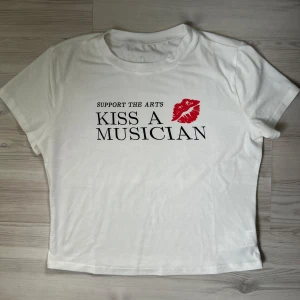 Vit graphic tee  - Säljer en graphic tee med texten 'Support the Arts, Kiss a Musician' och ett rött läppavtryck på framsidan. T-shirten är kortärmad och croppad, i storleken M. Materialet känns mjukt och bekvämt, idealiskt för vardagsbruk. Skriv gärna om du är intresserad innan du köper! 🫶