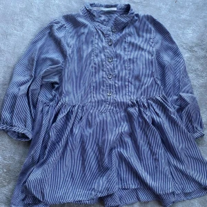 Blå och vit randig klänning från Custommade - Säljer en superfin blå och vit randig blus från Custommade. Den har en lös passform och är perfekt för både vardag och fest. Blusen har långa ärmar och knappar framtill. Materialet känns mjukt och bekvämt, och den har en lätt volangdetalj vid midjan som ger en söt touch. Perfekt för vår och sommar! 🌸