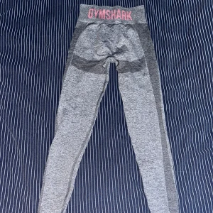 Gråa leggings från Gymshark - Säljer ett par gråa leggings från Gymshark i storlek S. De är högmidjade och har en tight passform som är perfekt för träning. Materialet är mjukt och stretchigt, vilket gör dem bekväma att ha på sig under hela träningspasset. Gymshark-loggan är tryckt i rosa på midjebandet och på sidan av benet. Leggingsen är i nyskick och redo för nya äventyr!